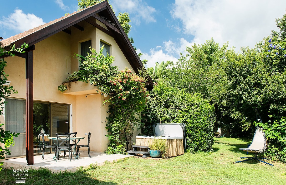 The exterior of the Bamboo House is highlighted by lush greenery and vibrant plants. A spacious patio features outdoor seating and a jacuzzi. The inviting lawn area offers various lounging spots under a bright sky, enhancing the connection with nature.