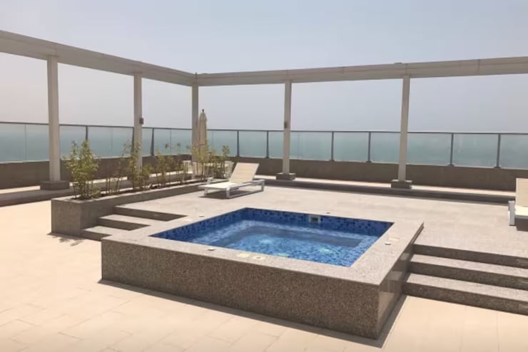An expansive rooftop terrace features a rectangular spa surrounded by a smooth stone deck. Sun loungers are arranged along the edges, with soft greenery providing a touch of nature. The backdrop includes unobstructed views of the sea and horizon.