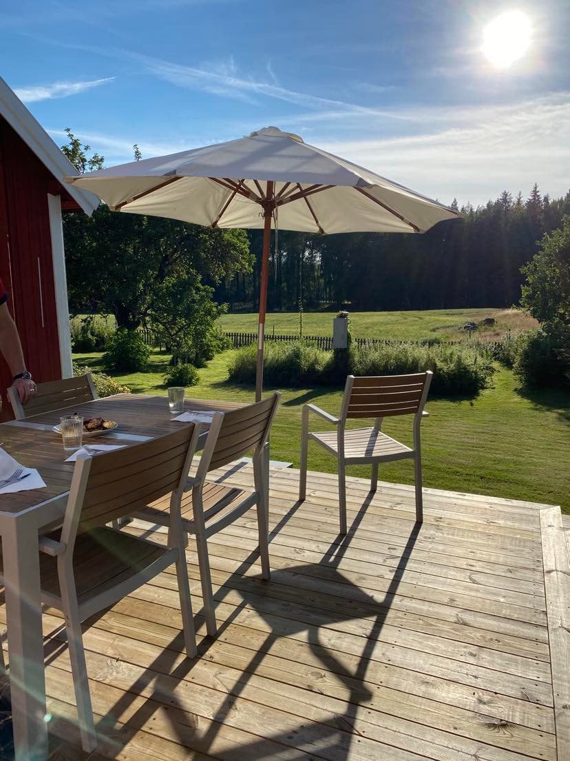 A wooden terrace features a dining table with several chairs beneath a large umbrella, providing shade. Lush greenery extends into the distance, with trees lining the horizon. Natural sunlight casts soft shadows, enhancing the peaceful outdoor setting.