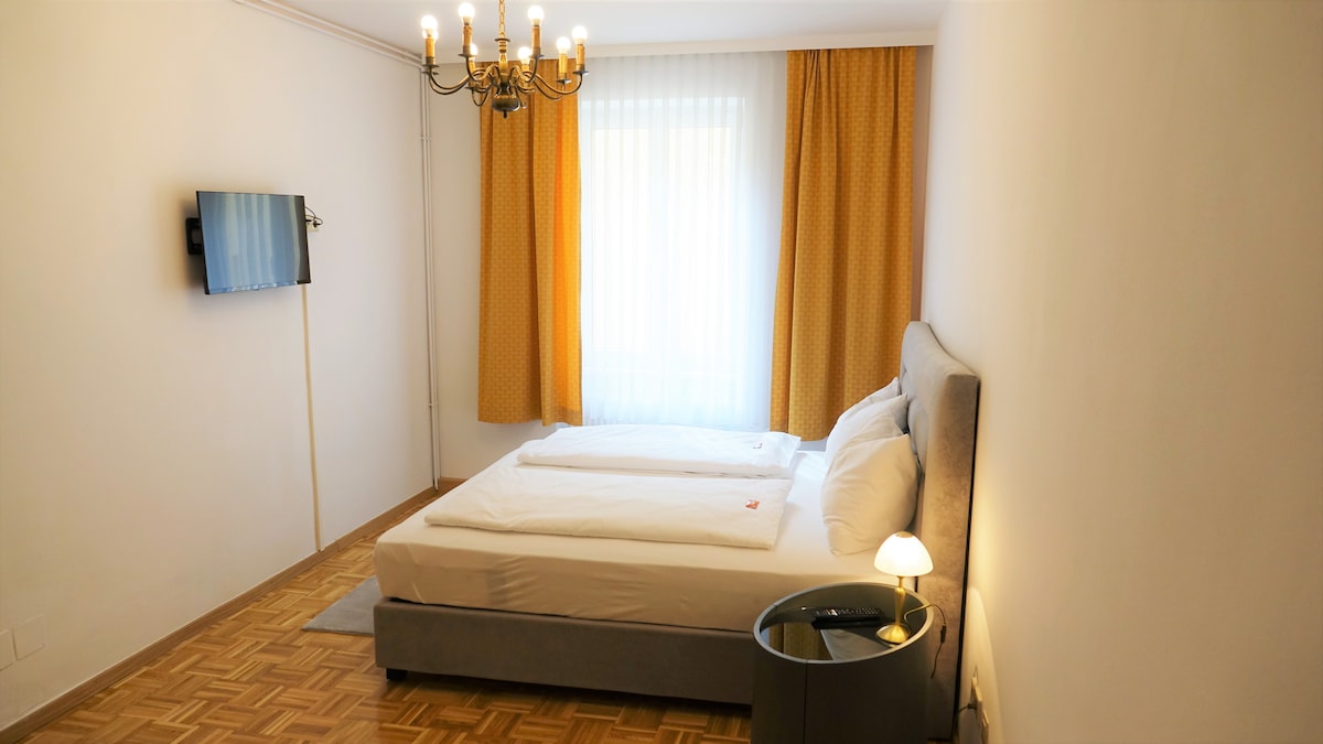 The sleeping area features a comfortable bed with crisp linens and plush pillows. Soft yellow curtains frame a large window, allowing natural light to brighten the space. A minimalist bedside table holds a small lamp, and a flat-screen TV is mounted on the wall.