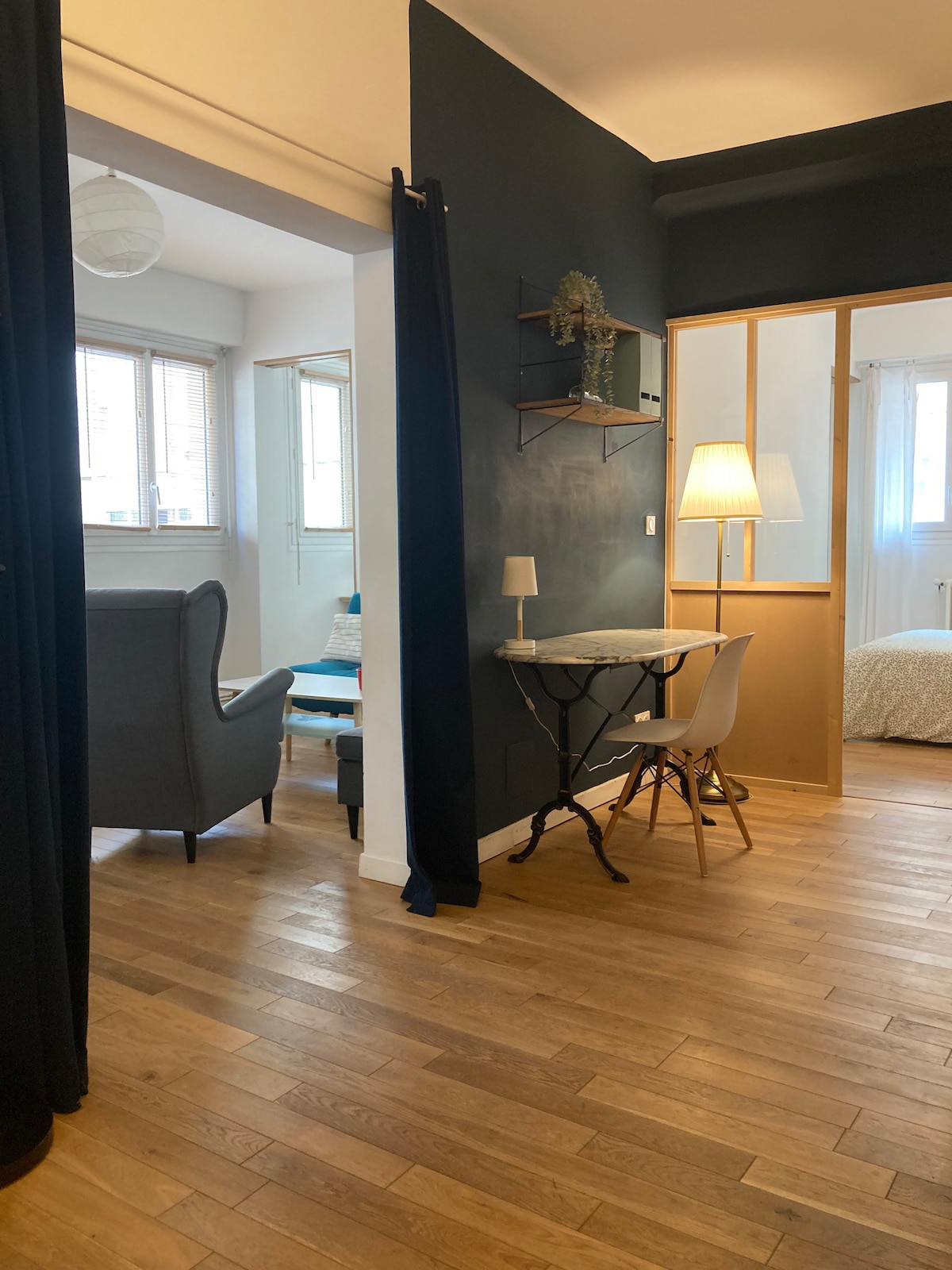A view into a well-lit apartment features a small workspace with a white chair and a black table. Natural light floods the area from windows framed in light wood. A stylish lamp stands beside a cozy seating area, enhancing the welcoming atmosphere.