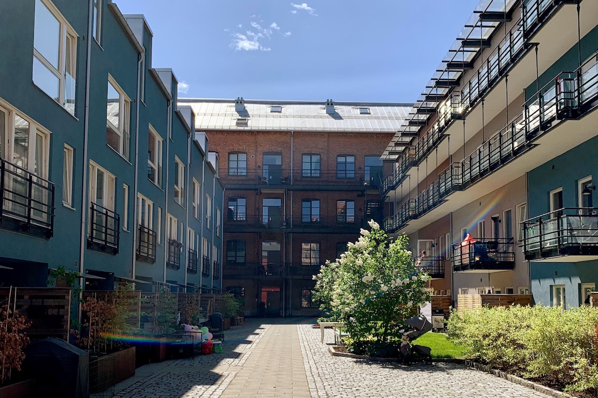 A serene courtyard is surrounded by modern buildings, featuring balconies on either side. Lush greenery and planted areas contribute to the tranquil atmosphere, while cobblestone pathways invite leisurely strolls through this cozy, sheltered space.