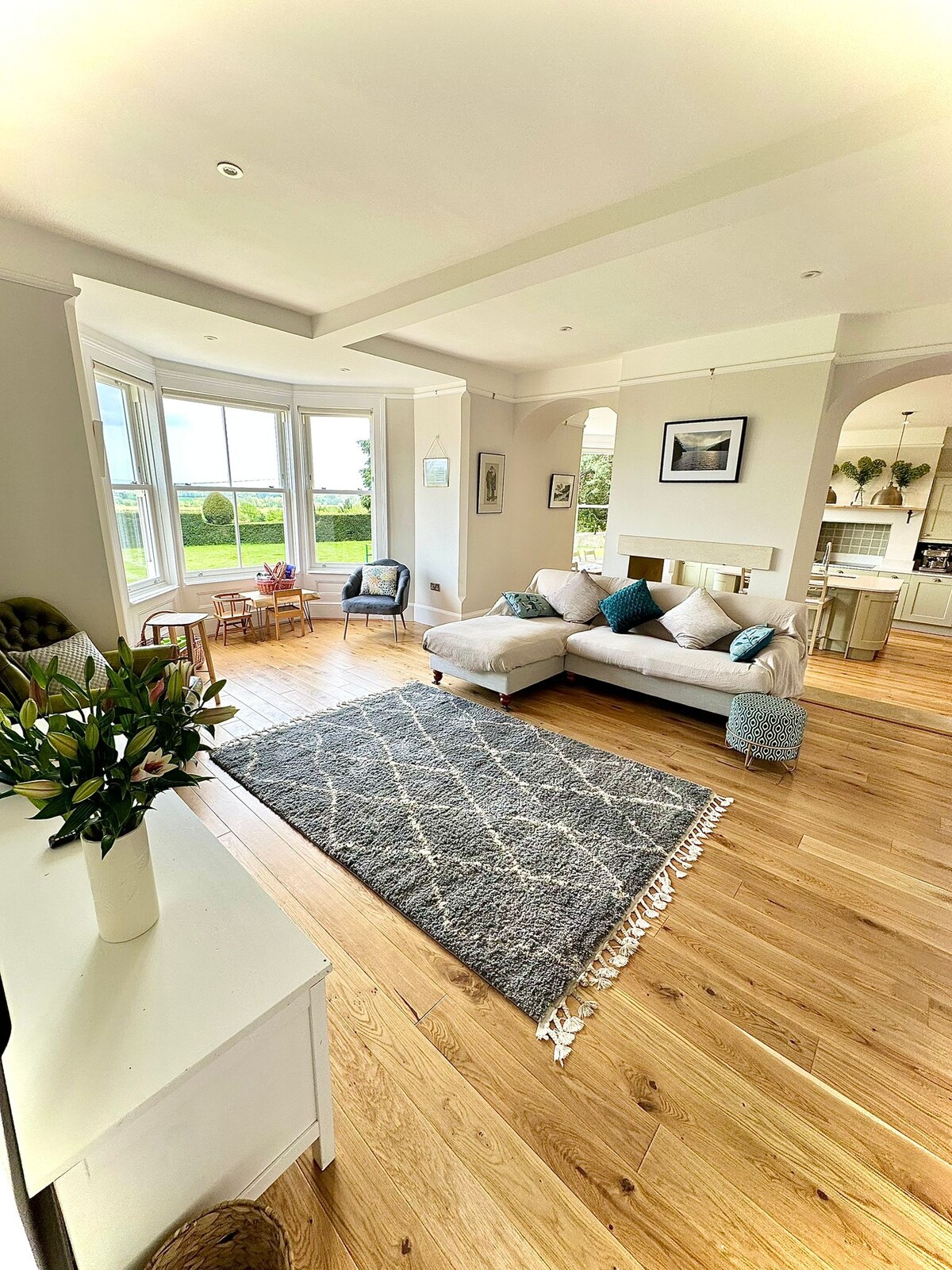 A bright, open living area features a large sectional sofa in neutral tones, accented with colorful cushions. A soft rug lies on polished wooden flooring. Large windows frame scenic views of the garden, allowing natural light to fill the space.