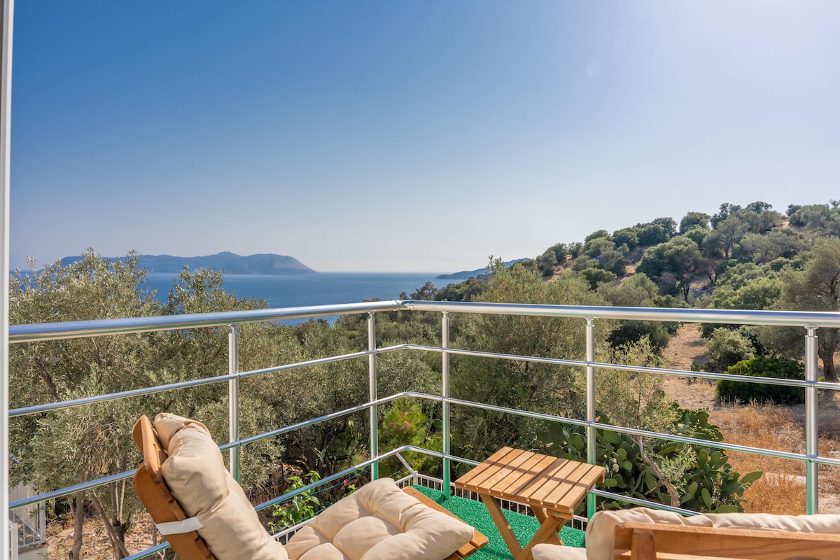 A cozy balcony area is furnished with a light-colored sofa and small wooden table. The expansive view includes rolling hills and the sea, under a clear blue sky, creating a sense of tranquility and connection with nature.