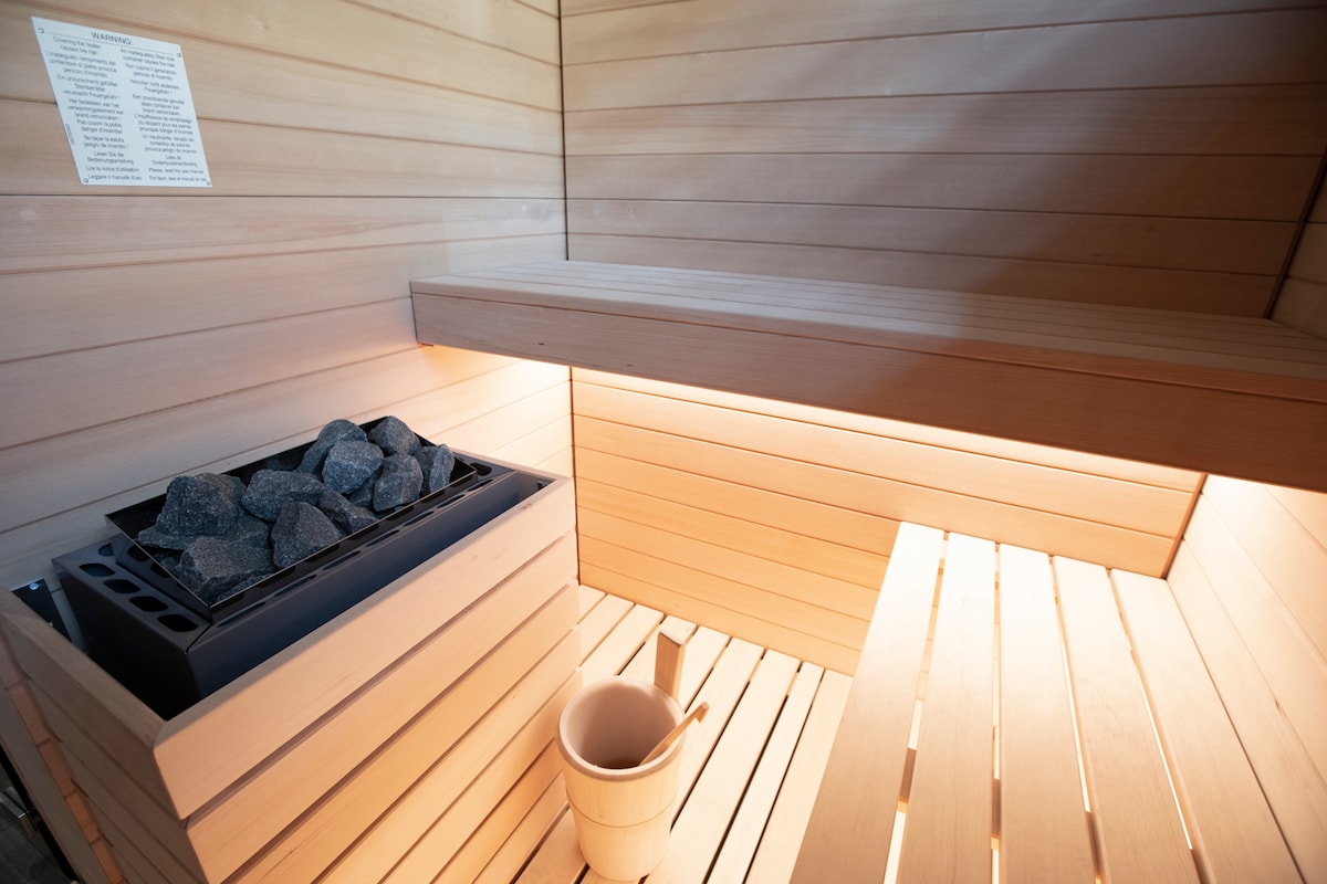 A wooden sauna interior features a stacked stone heater filled with dark stones. A light wooden bench runs along the walls, and a small wooden bucket is placed beside the heater. Soft illumination enhances the warm, natural tones of the wood.