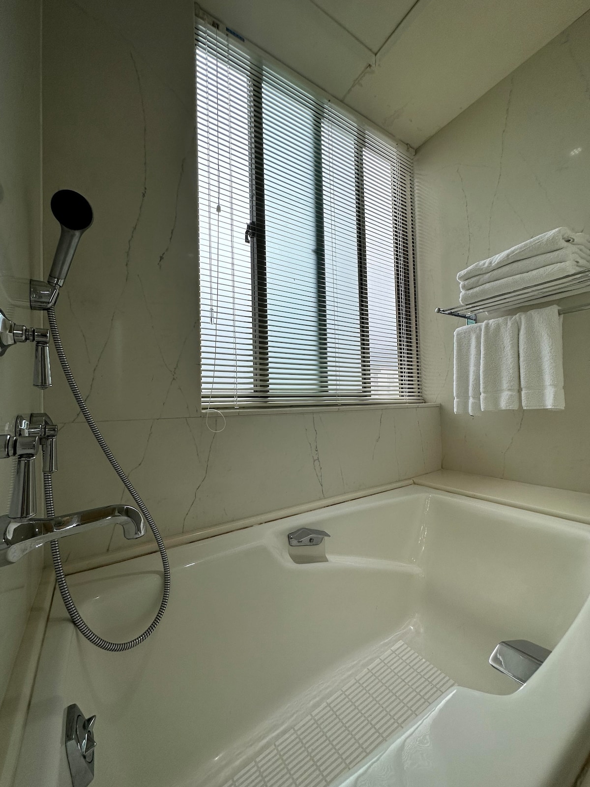 A modern bathroom features a spacious tub, equipped with a handheld showerhead. Natural light filters through a tall window adorned with blinds. Fresh white towels are neatly arranged on a wall-mounted rack nearby, adding to the clean and minimalistic design.
