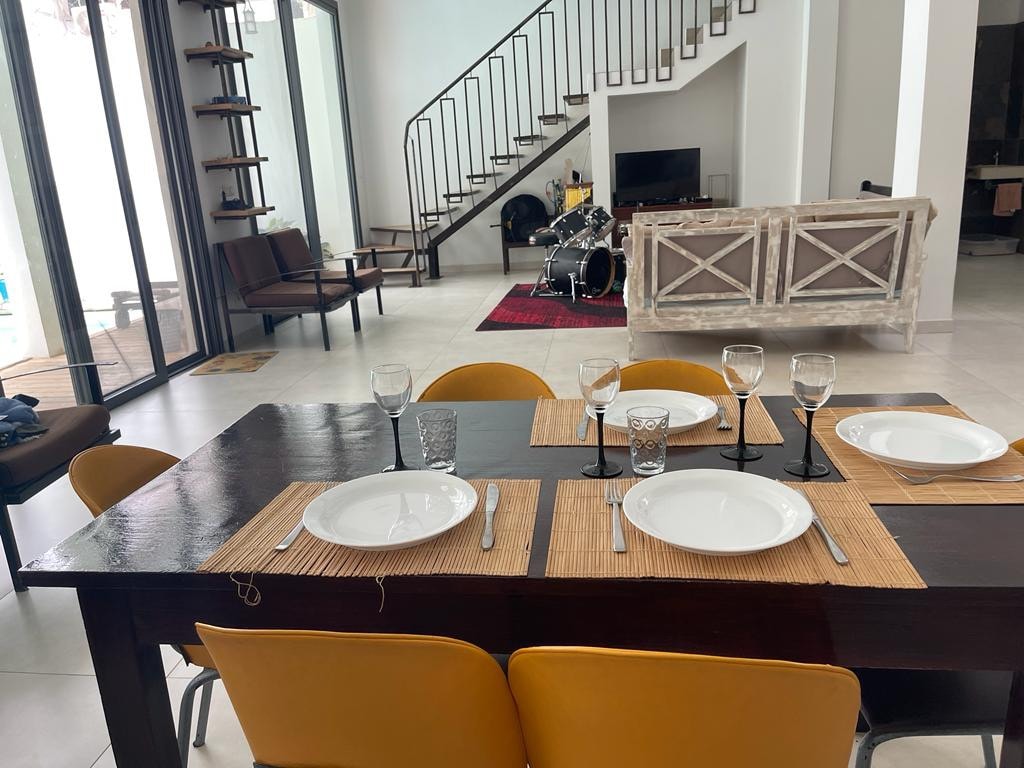 A dining table is set with white plates and glassware, surrounded by yellow chairs. A spacious living area is visible, featuring a staircase leading to an upper level. A glass door invites natural light, while a glimpse of a lounge area and decorative elements is present.