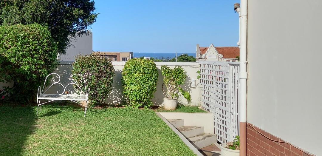A green lawn is bordered by well-maintained hedges and plants, leading to a view of the ocean in the distance. A white bench is positioned on the grass, providing a serene spot for relaxation. Steps lead from the lawn to a nearby pathway.