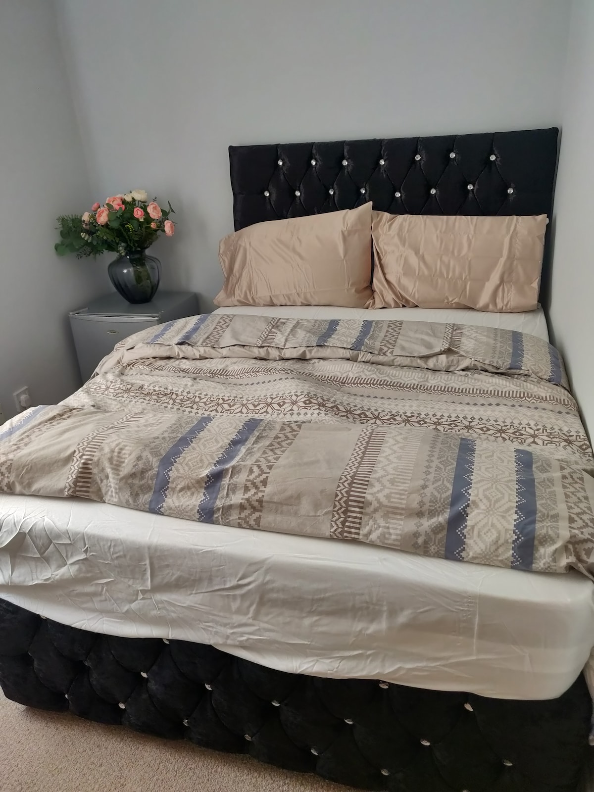 A cozy sleeping area is presented with a spacious bed featuring a tufted black headboard. Soft beige pillows rest on the bed, which is covered with a patterned blanket. A small nightstand holds a vase of fresh flowers, adding a touch of color to the room.