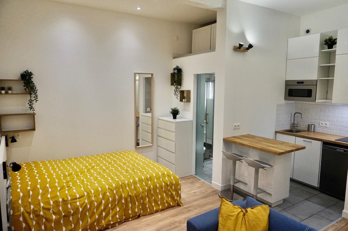 A studio apartment is depicted with a spacious yellow patterned bedspread covering a large bed. A minimalist kitchen area with a wooden countertop, modern appliances, and bar stools is visible. Natural light highlights the warm-toned wooden flooring and soft decor elements throughout the room.