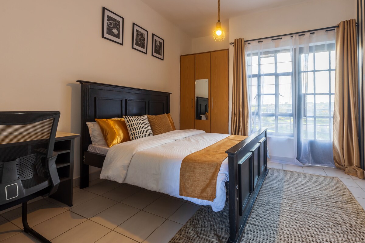 A well-appointed bedroom features a spacious bed dressed in white linens with decorative pillows. A desk is positioned nearby, complemented by a large window adorned with sheer curtains. Warm lighting highlights the wood accents and framed artwork on the walls.