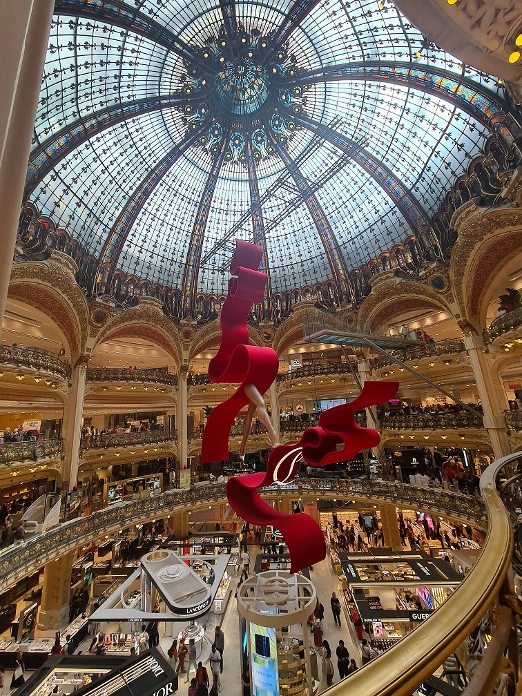 An intricate glass dome soars above a multi-level shopping arcade, adorned with elegant architectural details. A striking red sculpture hangs in the center, contrasting with the golden hues of the space, which is filled with visitors exploring various shops below.
