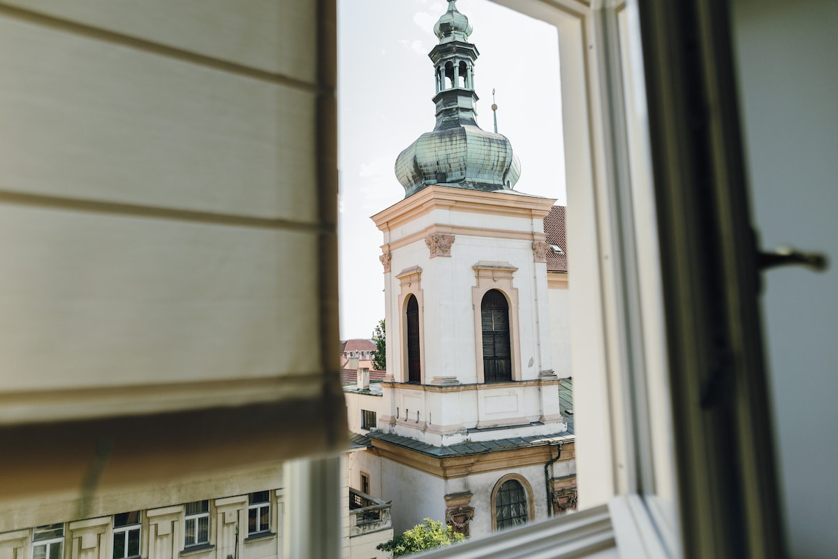 A view of the Baroque church of St. Ignatius of Loyola is framed by the window. The church features a prominent dome and intricate architectural details, showcasing its historical significance. Sunlight illuminates the structure, enhancing the visual appeal of this notable landmark in Prague.