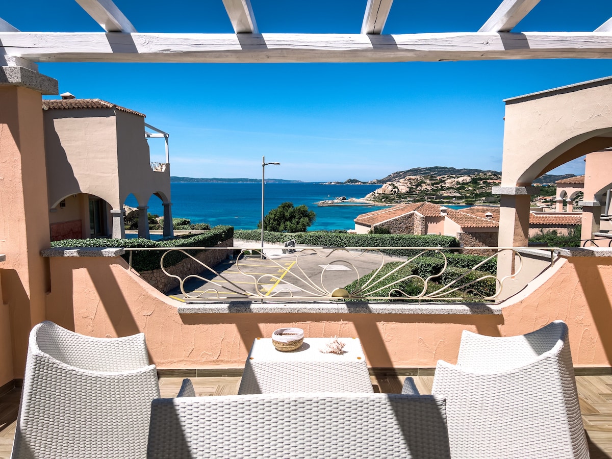 A terrace offers a serene view of the sea, framed by architectural arches. Comfortable white wicker seating is arranged around a small table, enhancing the outdoor space. The clear sky and distant coastline emphasize the tranquility of the setting.