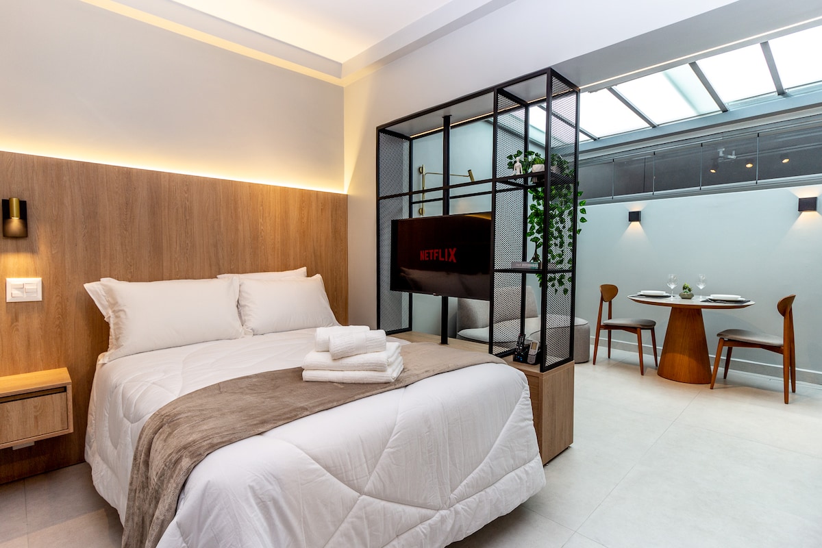 A modern room features a spacious bed with fresh linens and neatly arranged towels. A sleek TV is mounted on a decorative screen, highlighting entertainment options. A cozy dining area is visible, styled with wooden chairs and a round table, complemented by natural light from the glass ceiling.