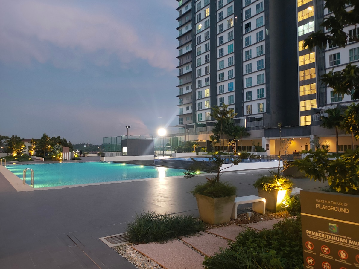 An outdoor area features a well-maintained swimming pool surrounded by greenery and modern buildings. Evening lighting enhances the ambiance, while a sign indicating the nearby playground can be seen. The scene reflects a calm atmosphere suitable for relaxation.