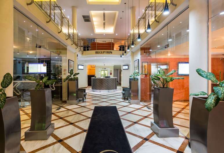 The modern reception area features a polished marble floor, bordered by geometric patterns. Tall glass walls provide visibility to welcoming greenery in planters. Bright lighting enhances the spacious atmosphere, with a service counter positioned in the background, complete with digital displays and seating arrangements.