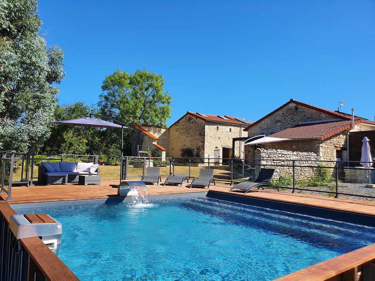 A refreshing swimming pool is framed by a wooden deck, featuring lounge chairs and shaded areas. Surrounding stone buildings and trees are visible in the background under a clear blue sky, providing a sense of calm in the outdoor space.