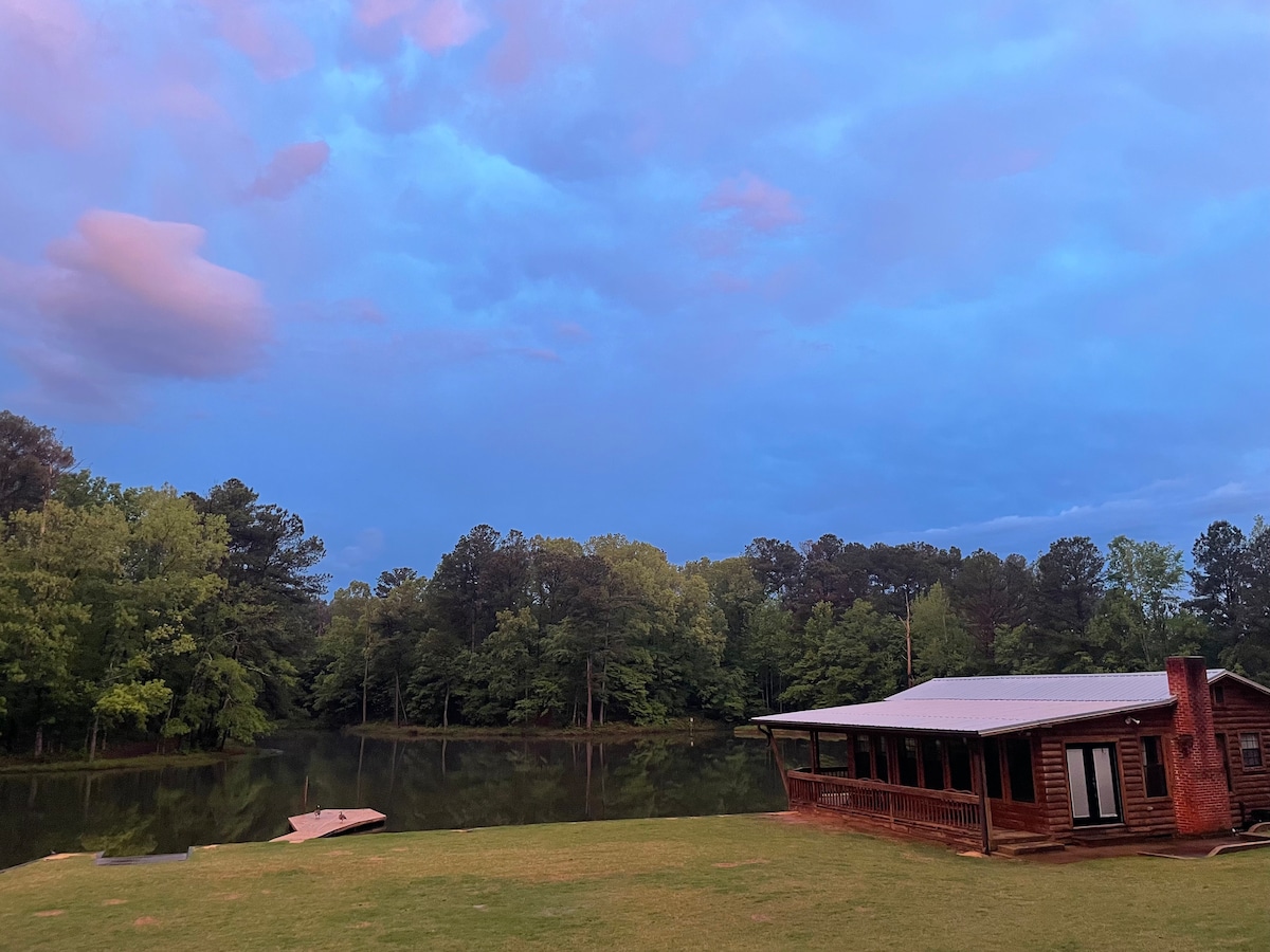 A cozy cabin is situated near a serene lake, surrounded by lush greenery. The building features large windows, allowing for views of the water. A small dock extends onto the lake, reflecting the calming hues of the sky during sunset.