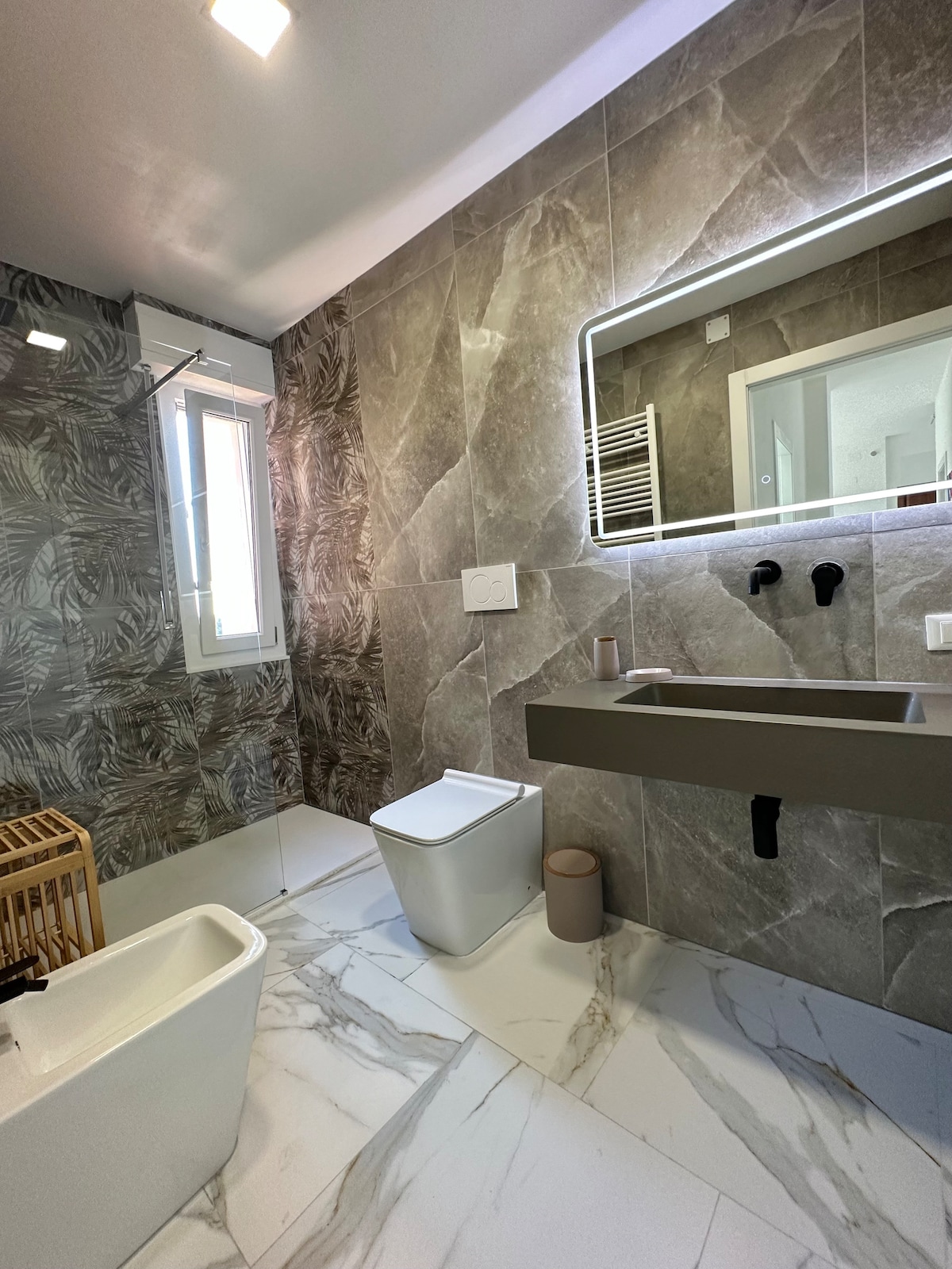 The bathroom features contemporary design elements, including a modern sink with a wall-mounted faucet and a shower area. Neutral-toned tiles adorn the walls and floor, while natural light enters through a window. A compact wooden storage unit adds functionality.