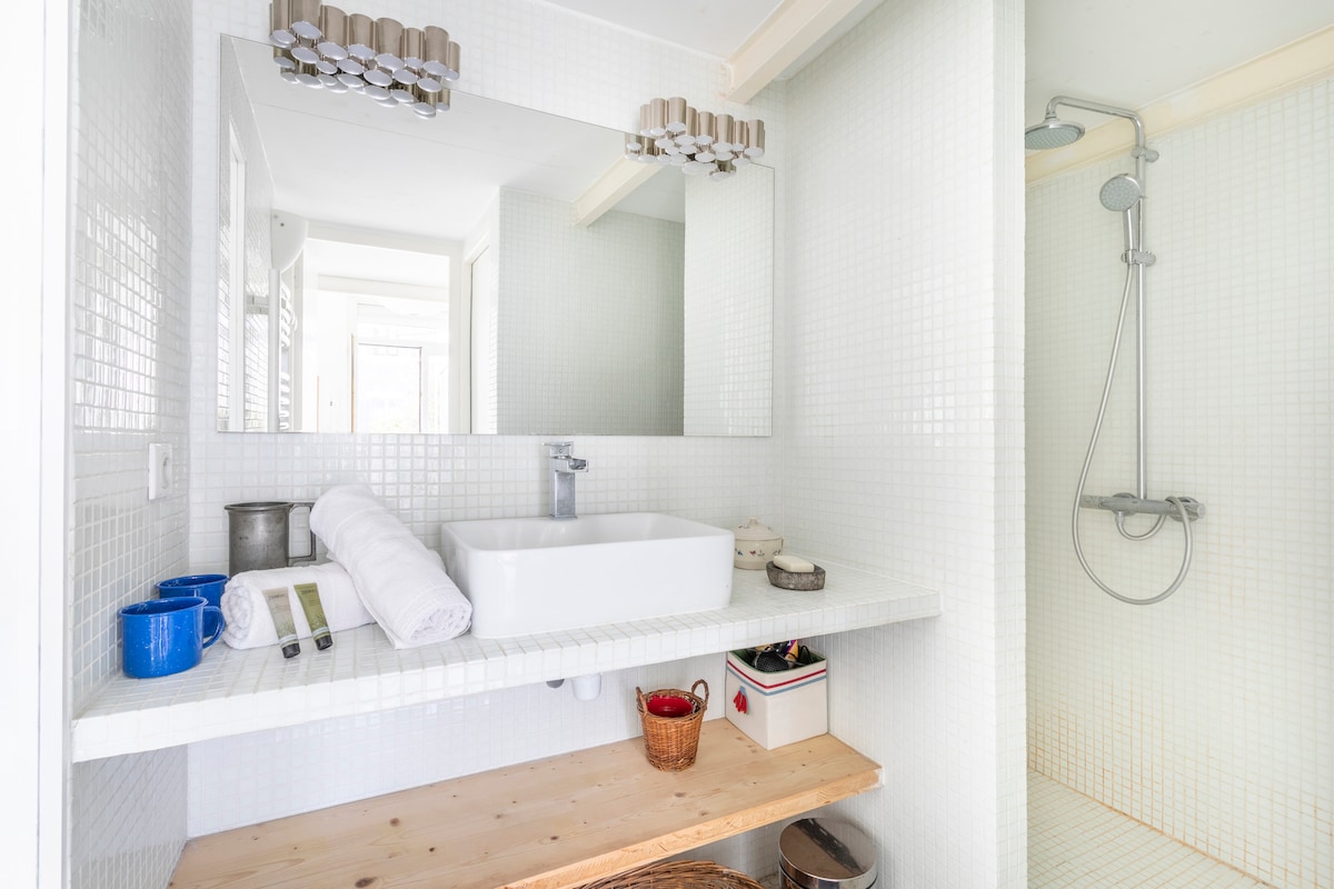 A bright and modern bathroom features a large wall-mounted mirror above a white basin sink. Soft towels are neatly arranged beside the sink, and various toiletries are placed on the countertop. The shower area is enclosed with tiled walls, enhancing a spacious feel.