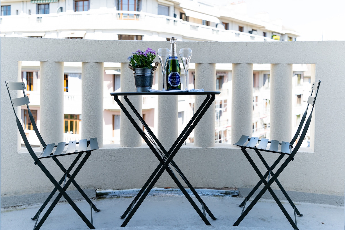 A balcony table is set with a bottle of champagne and two glasses, accompanied by a small potted plant. Two folding chairs frame the table, offering a comfortable spot to enjoy views of the surrounding area.
