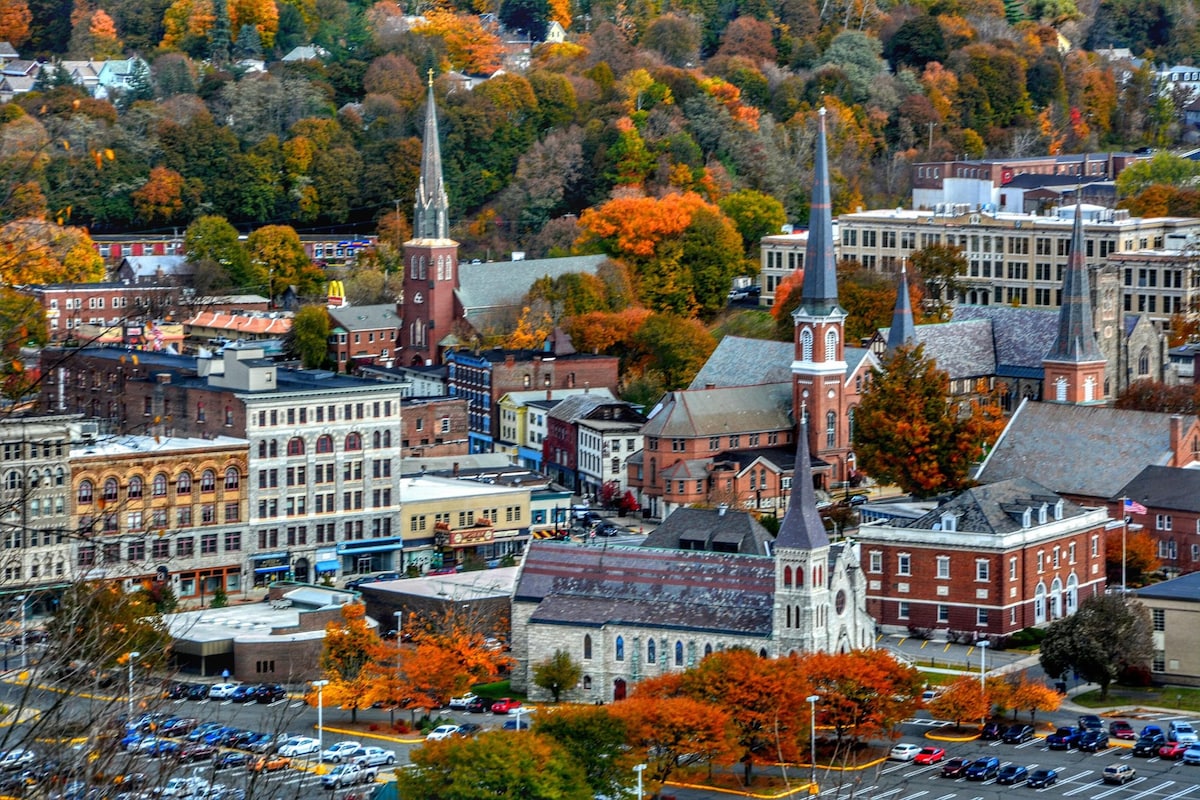 An aerial view captures the vibrant colors of autumn foliage surrounding downtown North Adams. The scene features historic buildings, steeples, and tree-lined streets, showcasing a mix of modern and classic architecture amidst the rich hues of fall.