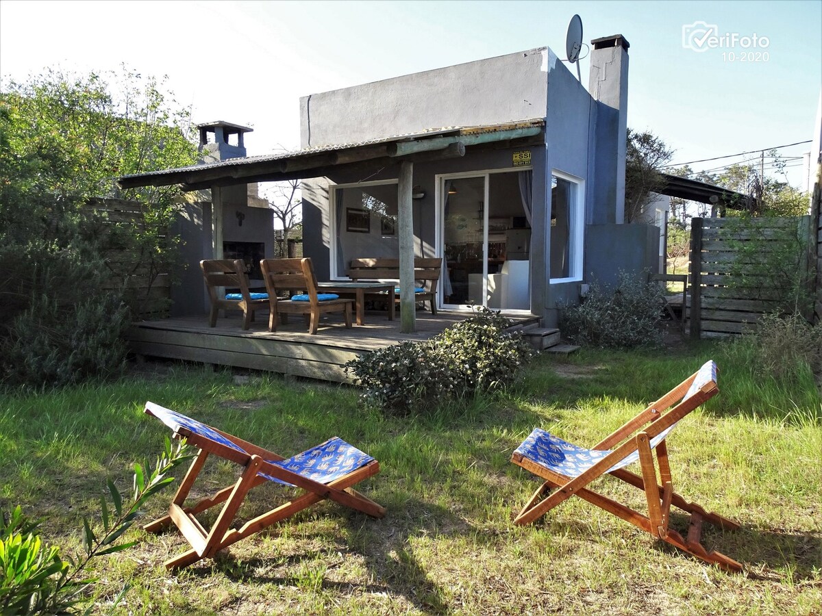 A simple, rustic cabin is viewed from the garden, featuring a spacious deck with outdoor seating. Two wooden lounge chairs are positioned in the foreground, facing the cabin. Large windows provide a glimpse into the interior, which is bathed in natural light.