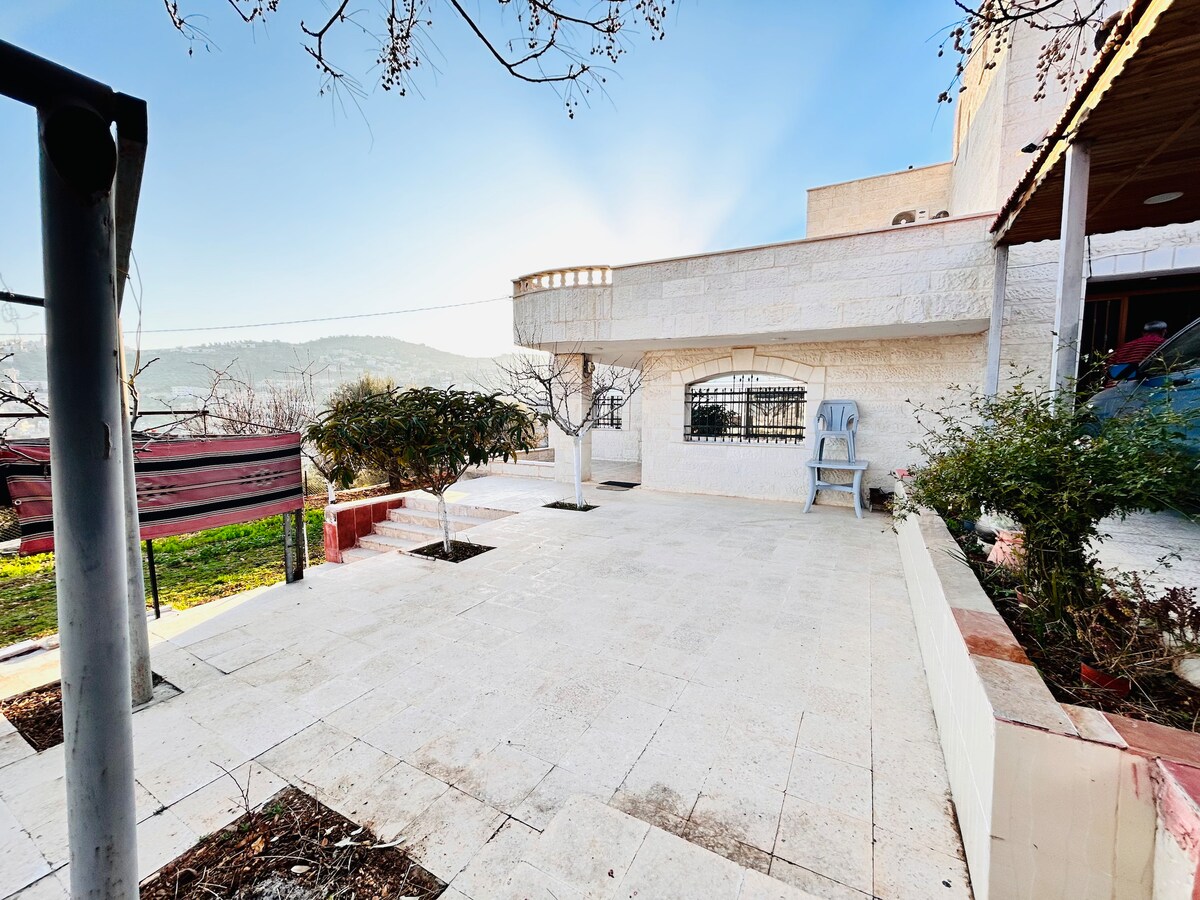 A spacious outdoor area is shown, featuring a patio with tiled flooring. A single chair is positioned nearby, and fruit trees are visible in the garden. The surrounding landscape includes gentle hills under a clear sky, enhancing the serene atmosphere of the space.