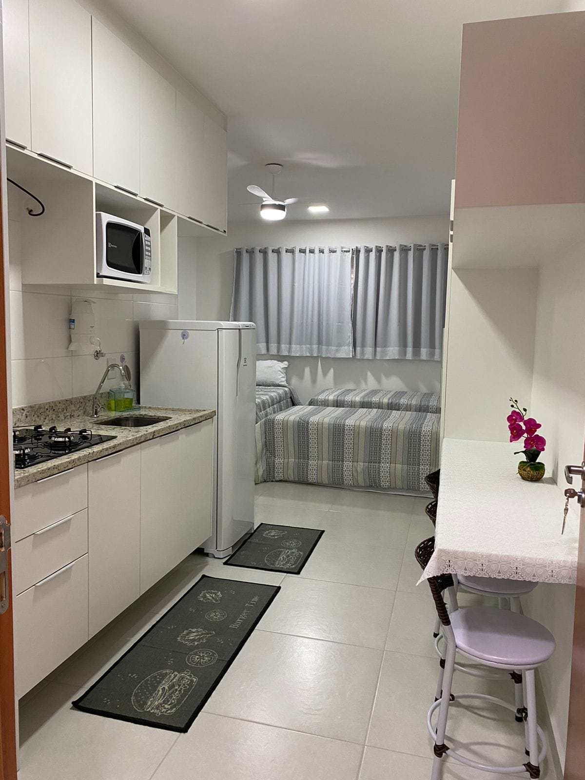 A compact kitchen area is visible, featuring modern cabinetry, a refrigerator, and a microwave above the counter. In the background, a sleeping area is shown with a neatly made bed, dressed in striped linens, and soft light filtering through closed blinds. A small dining space with two stools is positioned adjacent to the kitchen.