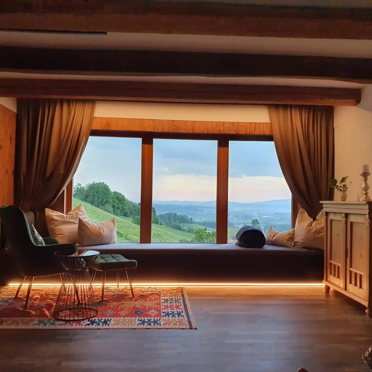 A cozy seating area features a large window overlooking the vineyards, framed by soft curtains. A rug with a colorful pattern lies on the floor, complementing a stylish armchair and a wire chair. The space is softly illuminated by warm lighting along the baseboard.