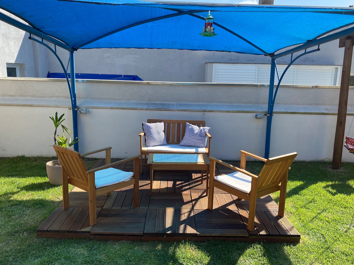 A shaded outdoor seating area features a wooden table surrounded by two armchairs on a wooden platform. Green grass surrounds the space, and a blue canopy provides cover from the sun. Decorative cushions are placed on the seating for added comfort.