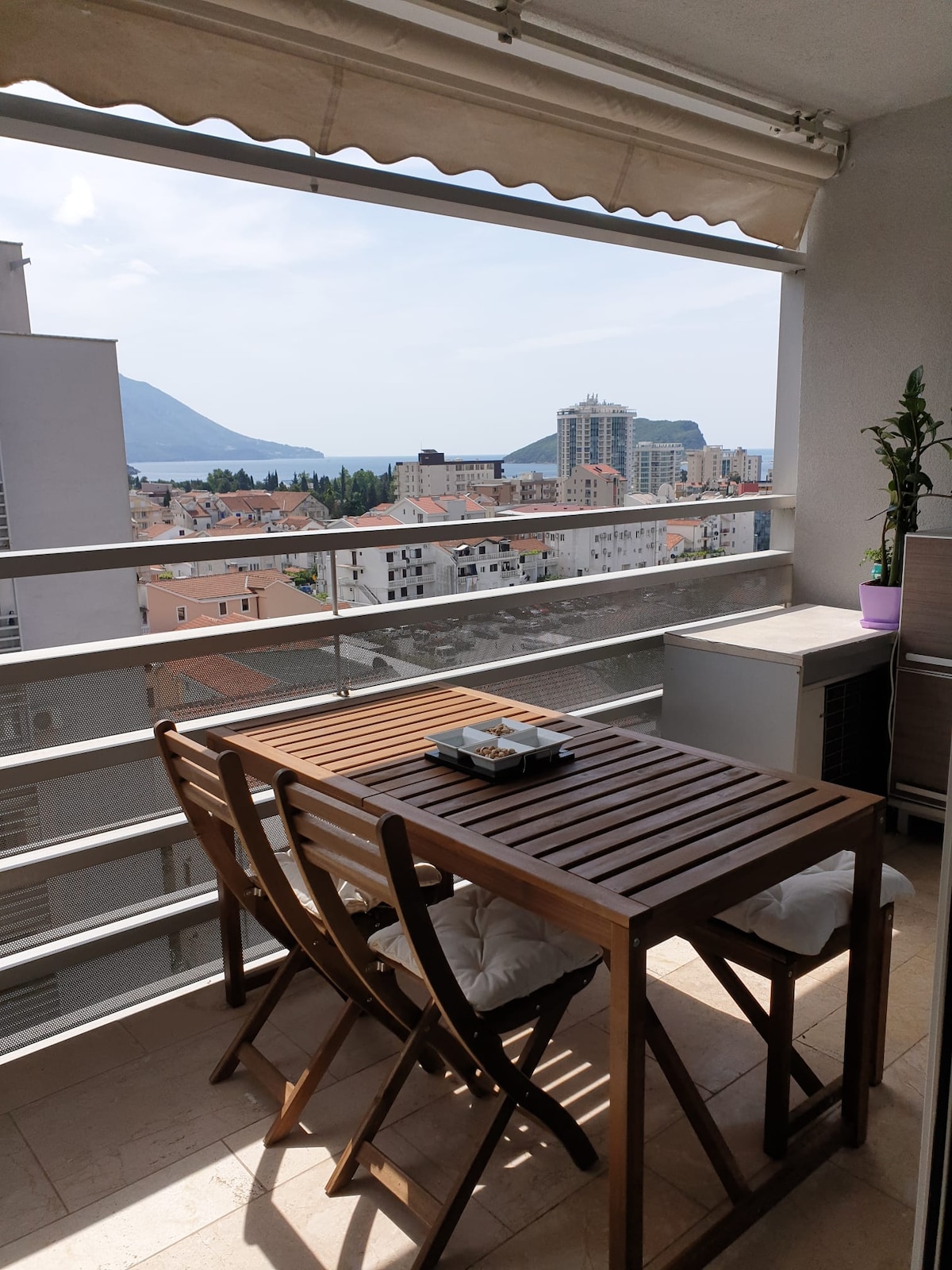 A balcony area is presented with a wooden table and two folding chairs, offering a spacious space for relaxation. The view showcases a scenic landscape, including distant mountains and the sea, with buildings seen below, framed by clear skies.