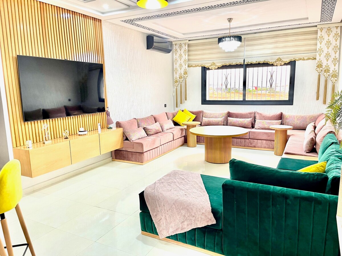 A spacious living area features a large sectional sofa with soft pink upholstery and green accents. A round wooden coffee table is centered in the space, complemented by a modern flat-screen TV mounted on the wall. Natural light filters in through secured windows adorned with elegant drapes.