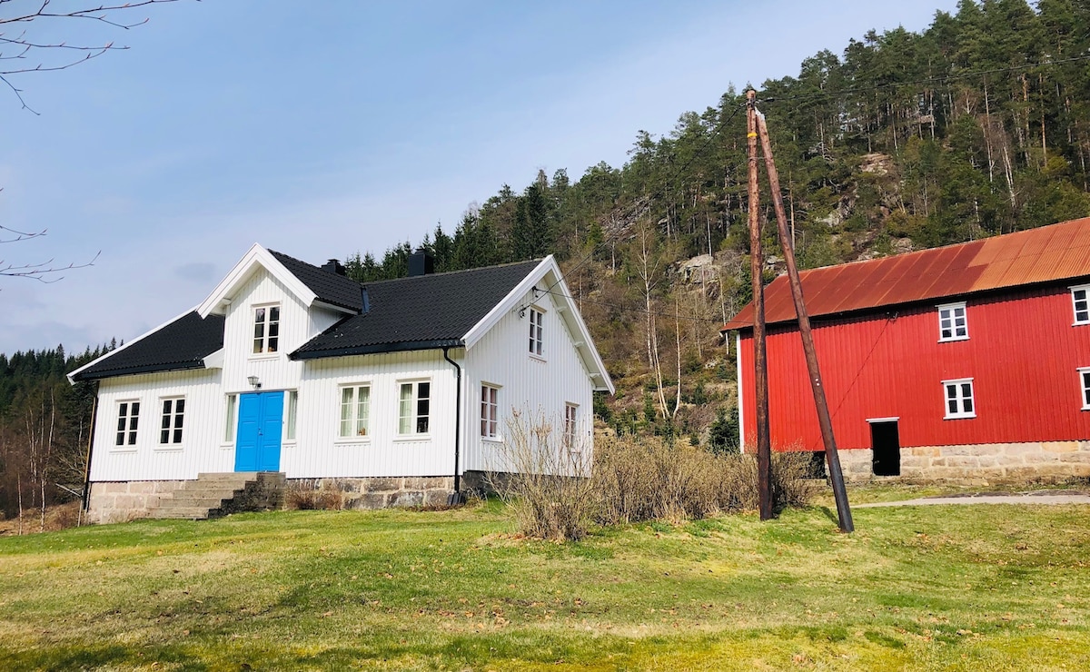 A classic white farmhouse with a blue door is set against a backdrop of green trees and rocky hills. A charming red barn stands nearby, completing the rustic landscape. A spacious lawn surrounds the house, providing an open and serene atmosphere.