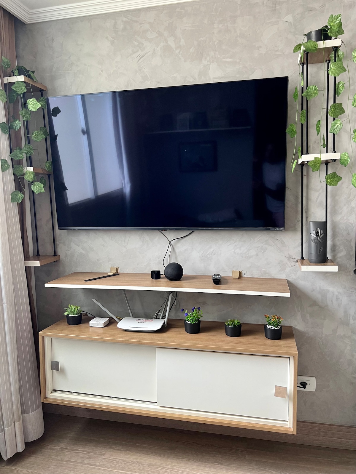 A modern entertainment unit is displayed against a textured wall. A large television is mounted above a sleek shelf, designed with plants and decorative items. A minimalist lower unit features sliding doors and additional plants, enhancing the space's contemporary aesthetic.