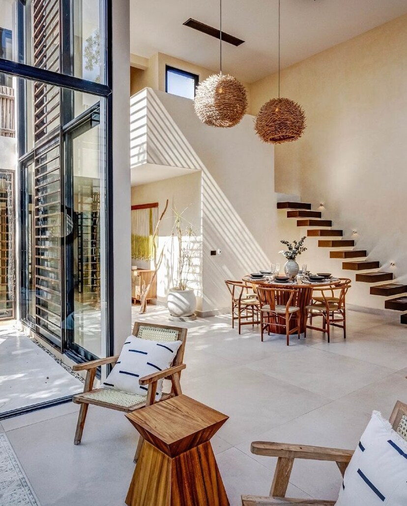 An airy living space features natural light pouring in through floor-to-ceiling glass windows. A dining table is set for six, surrounded by wooden chairs. A cozy seating area includes two armchairs with patterned cushions and a wooden side table, complemented by a staircase with modern steps.