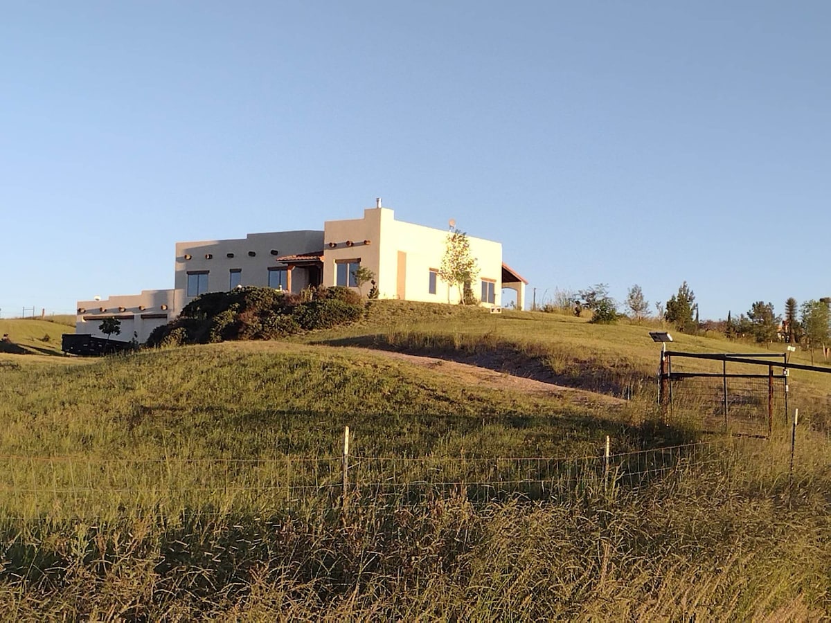 A luxury home situated on a hilltop is depicted, surrounded by rolling hills. The structure features a modern design with expansive windows and outdoor porches, blending harmoniously with the natural landscape. The property is set on five fenced acres, enhancing privacy and openness.