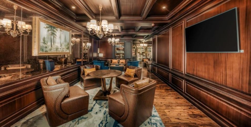 An elegant lounge is showcased with dark wood paneling and soft lighting from stylish chandeliers. Comfortable leather chairs surround a round table atop a textured rug. A large painting adorns one wall, and a modern television is mounted nearby, enhancing the inviting ambiance.