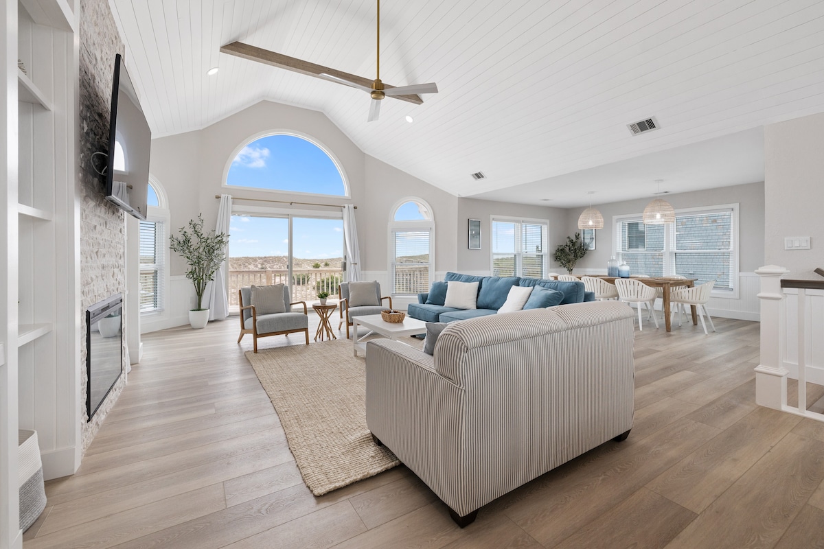 An open living area is highlighted by a cathedral ship lap ceiling and large arched windows, allowing natural light to fill the space. A cozy sectional couch faces a wall-mounted TV and an electric fireplace. A dining table and soft area rug add warmth to the environment.