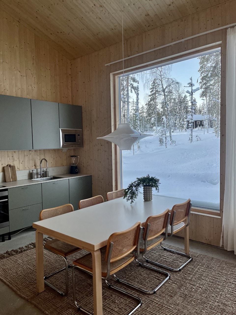 A cozy dining area features a spacious white table surrounded by six wooden chairs. Large windows frame a winter landscape with snow-covered trees outside. Neutral tones of the wood paneling and cabinetry complement the minimalist design.