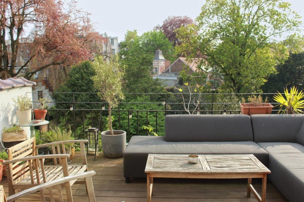 A spacious terrace is presented, featuring a comfortable grey seating area and a wooden coffee table. Potted plants are arranged around the space, complementing the lush greenery of trees visible in the backdrop, providing a serene view of the surrounding neighborhood.
