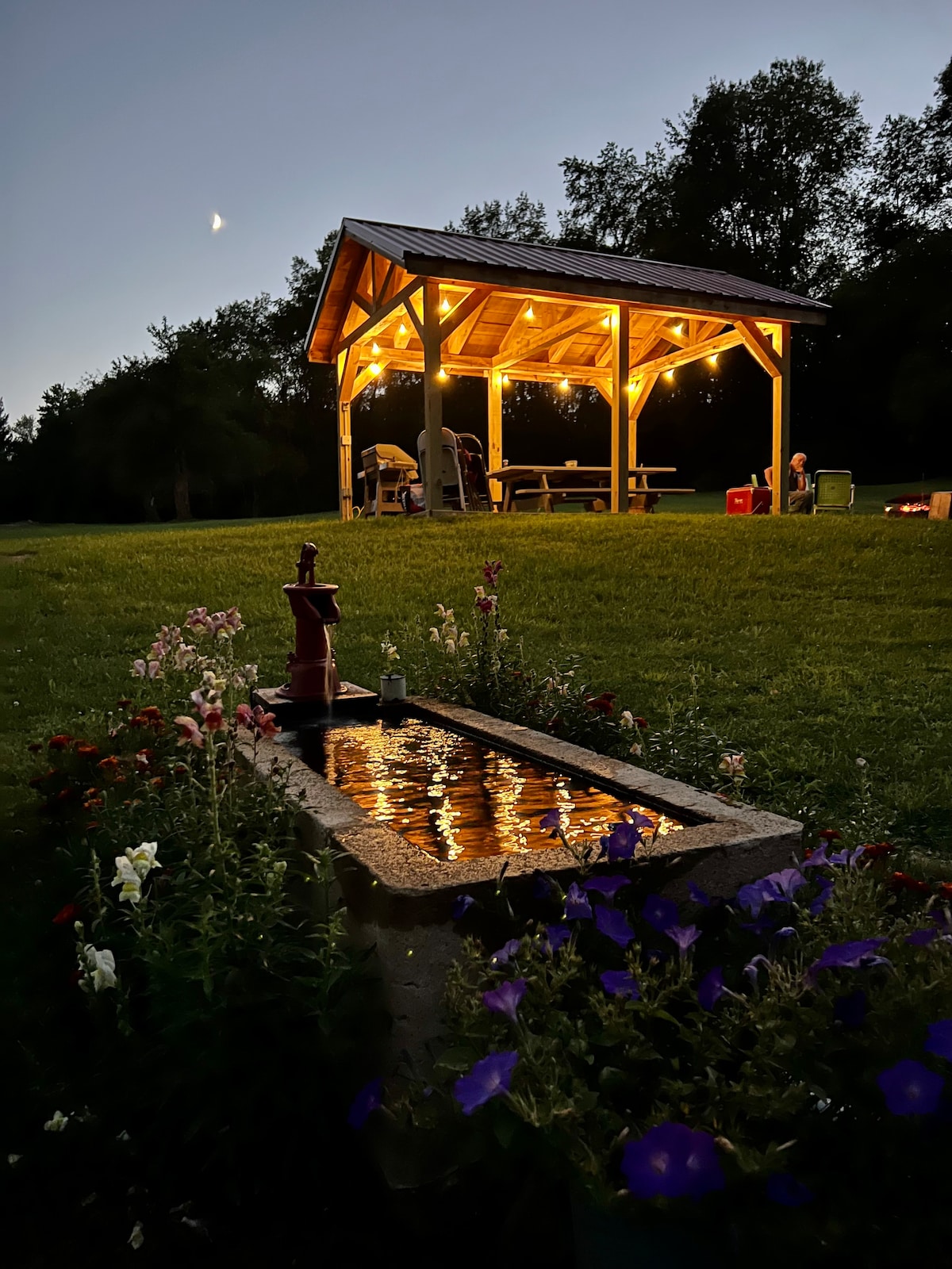 A pavilion is illuminated by soft lights at dusk, creating a welcoming gathering space. A flowerbed with various blooms borders a rustic stone water feature, its surface reflecting the evening glow. Seating is available under the pavilion, inviting relaxation and socializing.