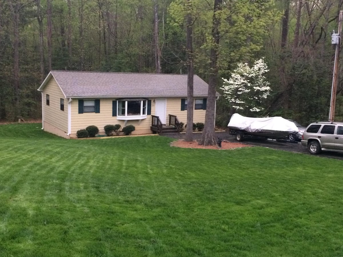 A single-story home with a light yellow exterior is set amidst lush greenery. The front features a small wooden deck with steps leading up, surrounded by manicured grass and neatly trimmed shrubs. A covered boat is parked beside the house on a gravel area.