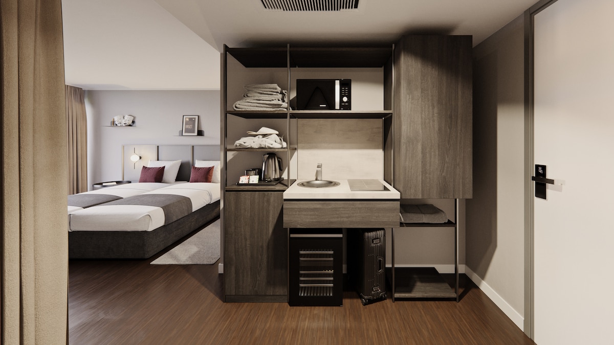 A kitchenette area is equipped with modern appliances including a microwave, kettle, and refrigerator. Neatly arranged shelves hold towels and kitchenware. The background features a spacious bed with neutral bedding, complemented by soft lighting from the nearby window.