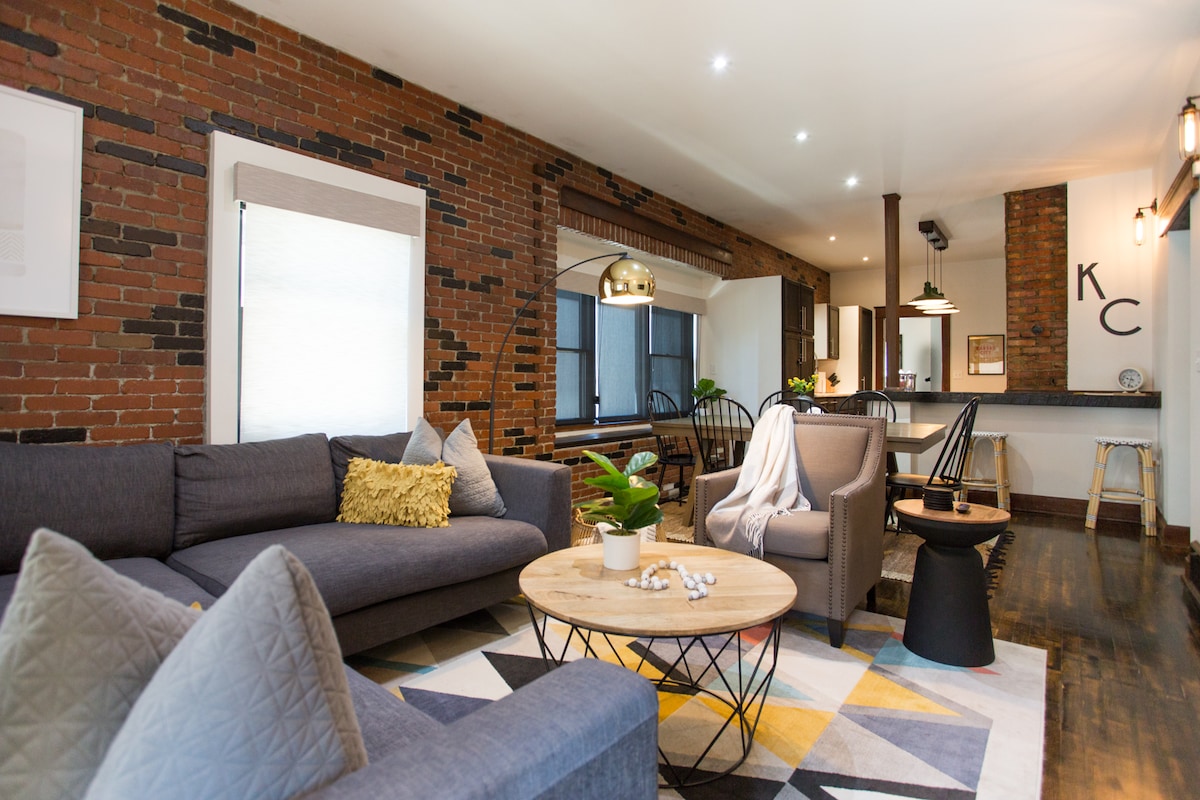 A spacious living area features a mix of modern and rustic elements, including a cozy sectional sofa, accent chairs, and a round wooden coffee table. A geometric area rug enhances the room, while exposed brick walls add character. Soft lighting creates a welcoming atmosphere.