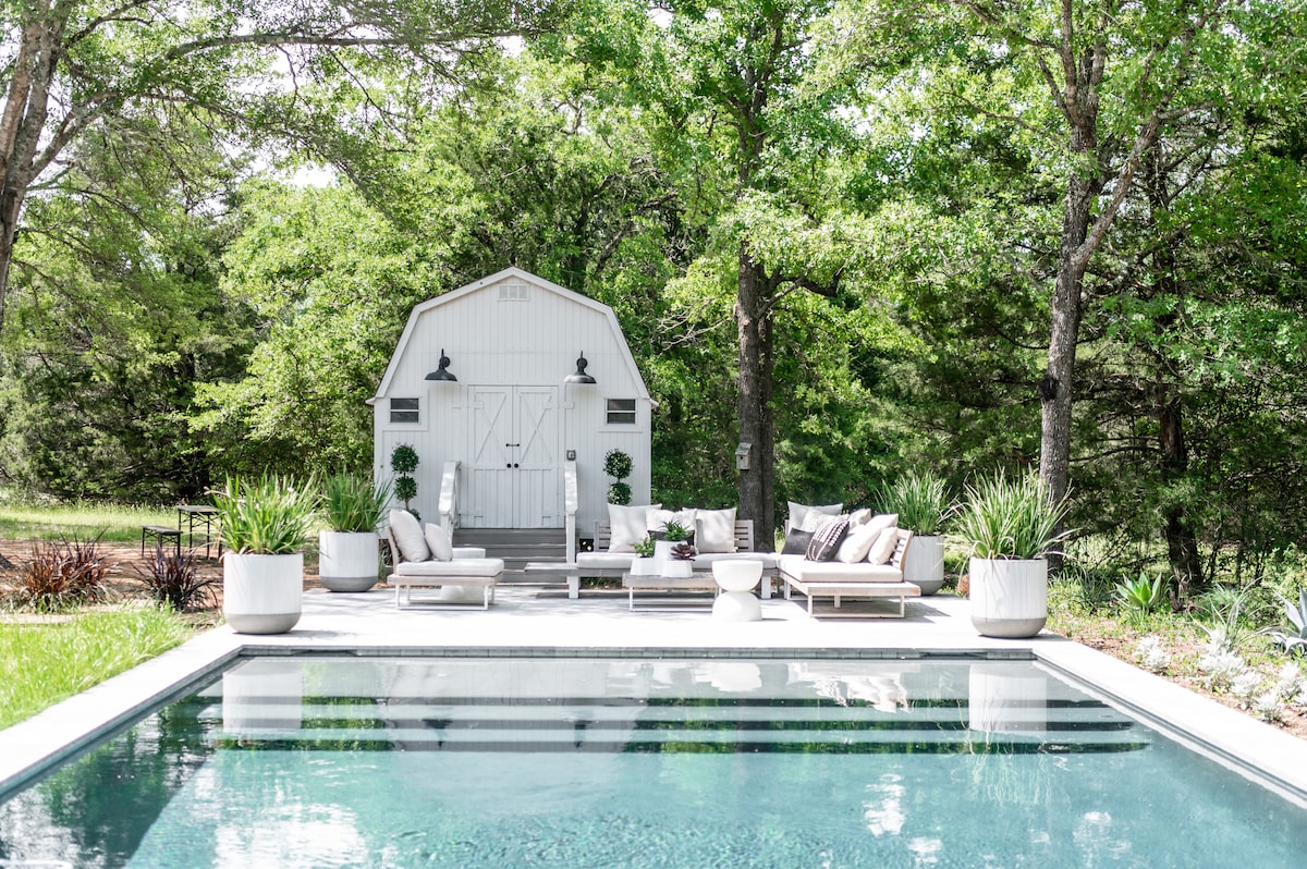 A serene pool area is surrounded by lush greenery, featuring a modern-style pool with clear water. Comfortable seating arrangements with neutral cushions face the pool, accompanied by large planters. A charming building is visible in the background, blending harmoniously with the natural landscape.