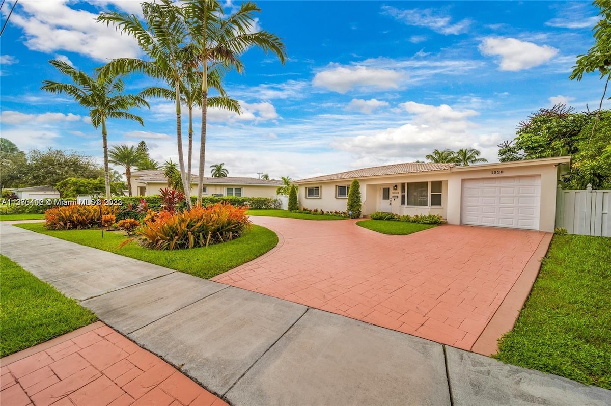 A well-maintained property is set within a landscaped yard featuring vibrant tropical plants. A wide, paved driveway leads to a one-car garage, while palm trees add a touch of greenery against a clear blue sky. The exterior is characterized by a light-colored facade.