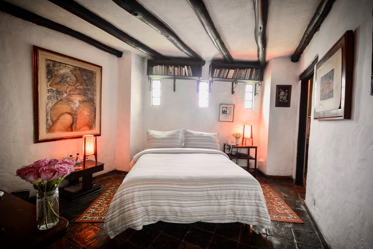 An intimate sleeping area showcases a double bed with lightly striped bedding, flanked by two lamps casting a soft glow. Nearby, an area rug complements the dark floor, while shelves display books. Artworks adorn the walls, enhancing a cozy ambiance.