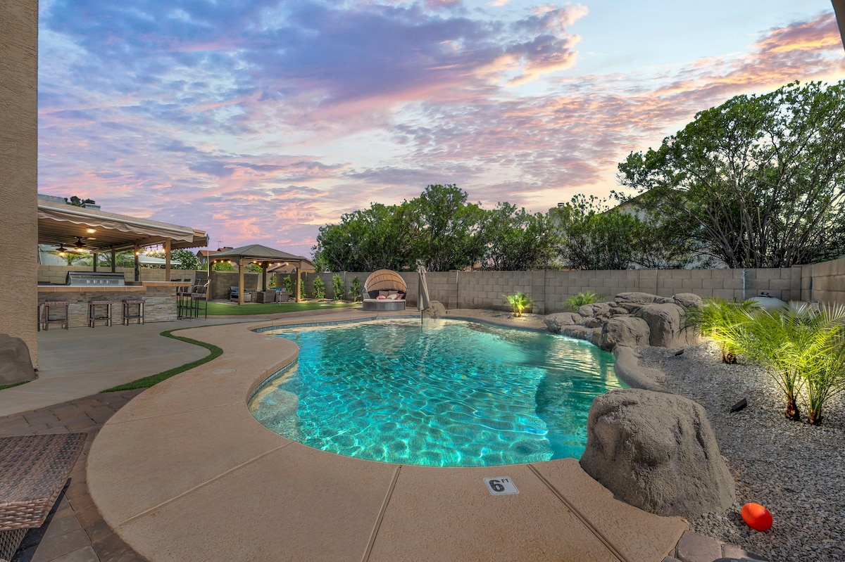 A serene backyard features a sparkling swimming pool surrounded by landscaped greenery. Two gazebos provide shaded seating areas, while natural stone accents enhance the outdoor aesthetic. A soft sunset sky casts warm hues over the tranquil scene, inviting relaxation and enjoyment.